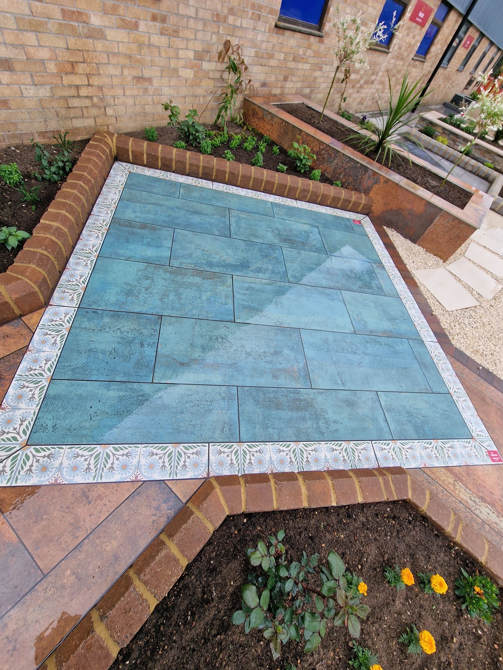Blue green porcelain patio with a patterned tile border and brick raised beds